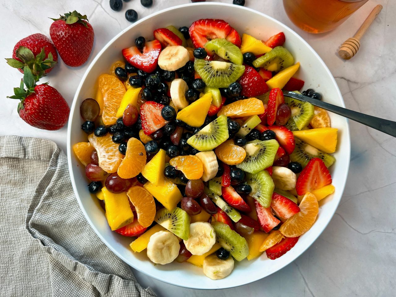 Fruit Salad in a white bowl with a black spoon and honey on the side