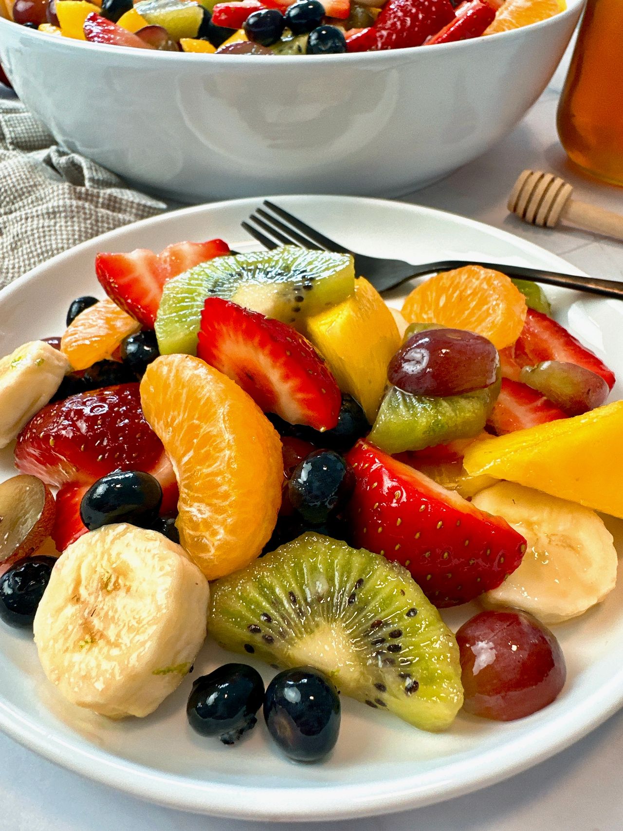 Fruit Salad in a white bowl with a black spoon and honey on the side