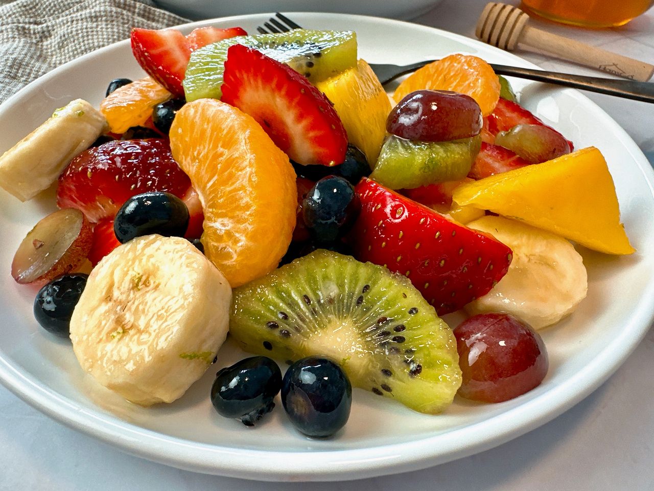 Fruit Salad in a white bowl with a black spoon and honey on the side