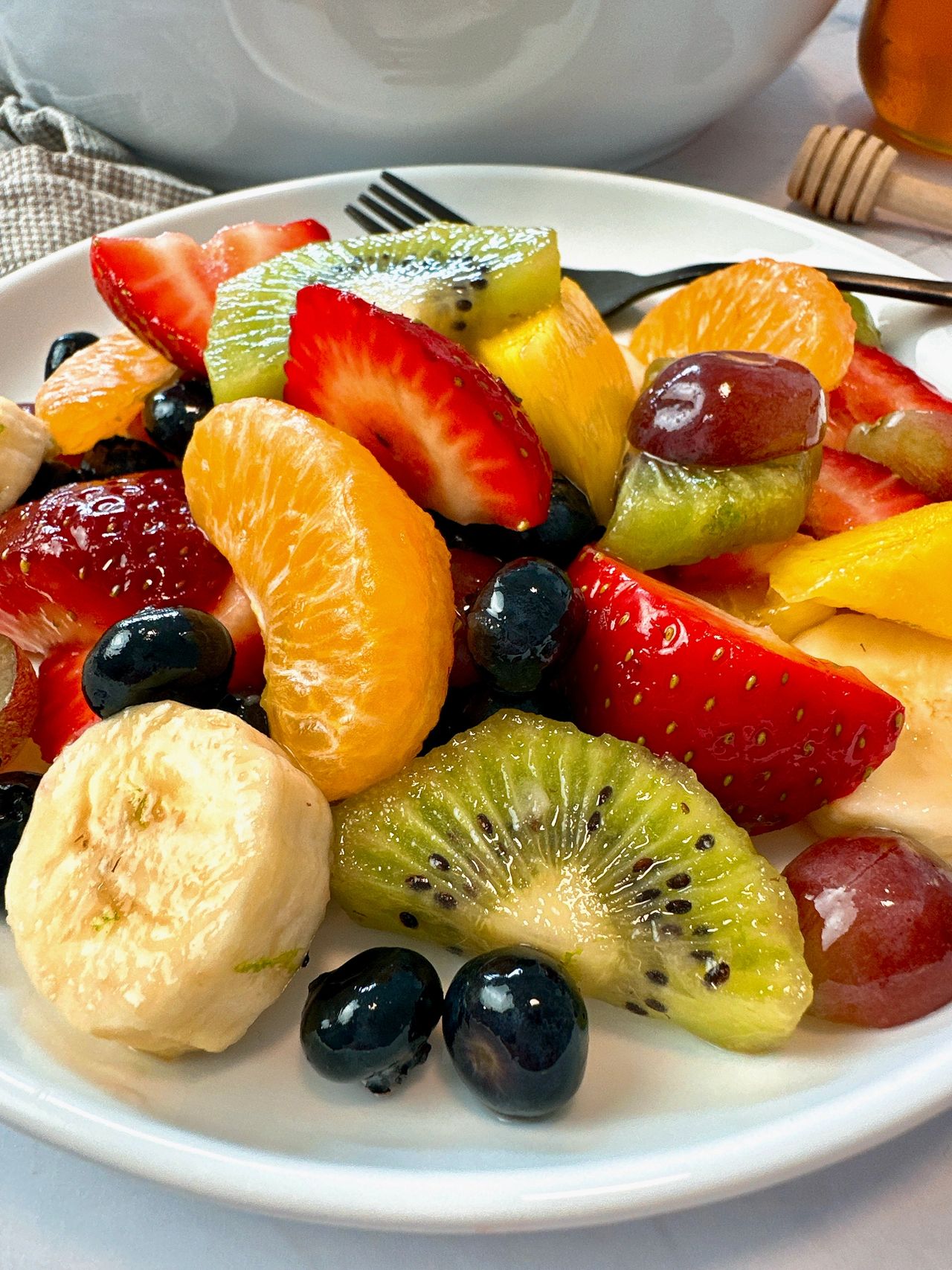 Fruit Salad in a white bowl with a black spoon and honey on the side