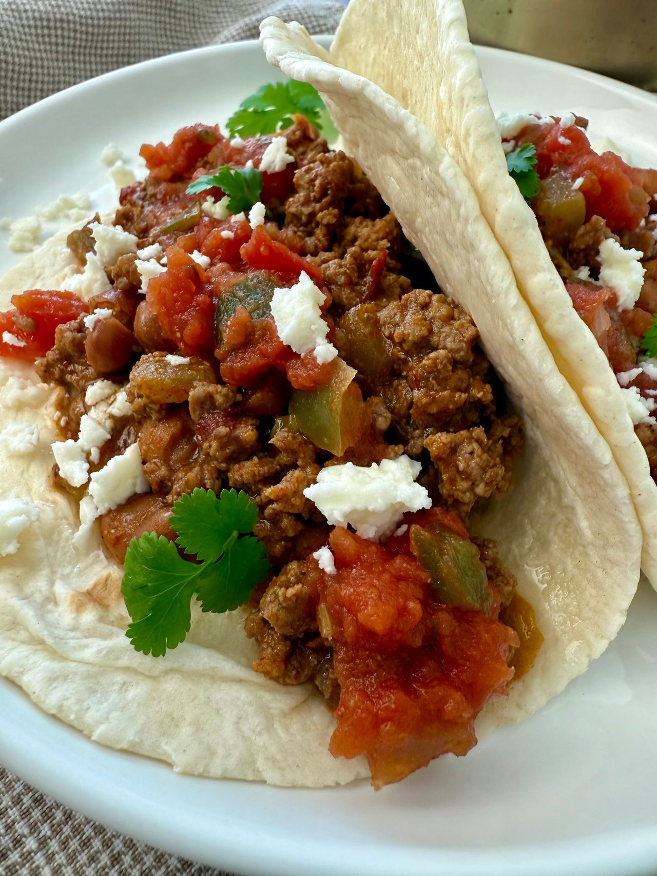 ground beef and bean soft tacos on a white plate with crumbled cheese garnish
