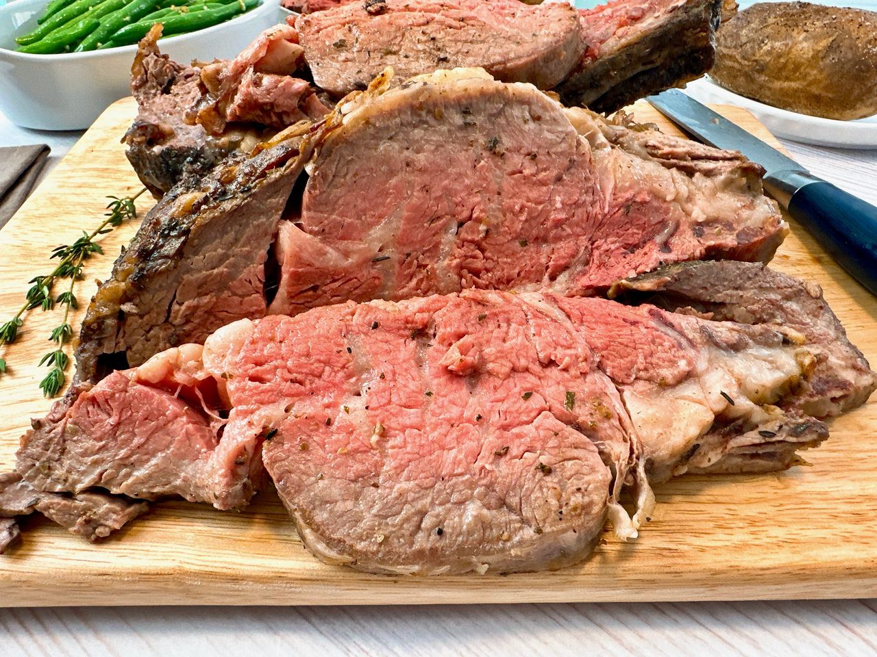slices of prime rib on a cutting board with a baked potato and green beans