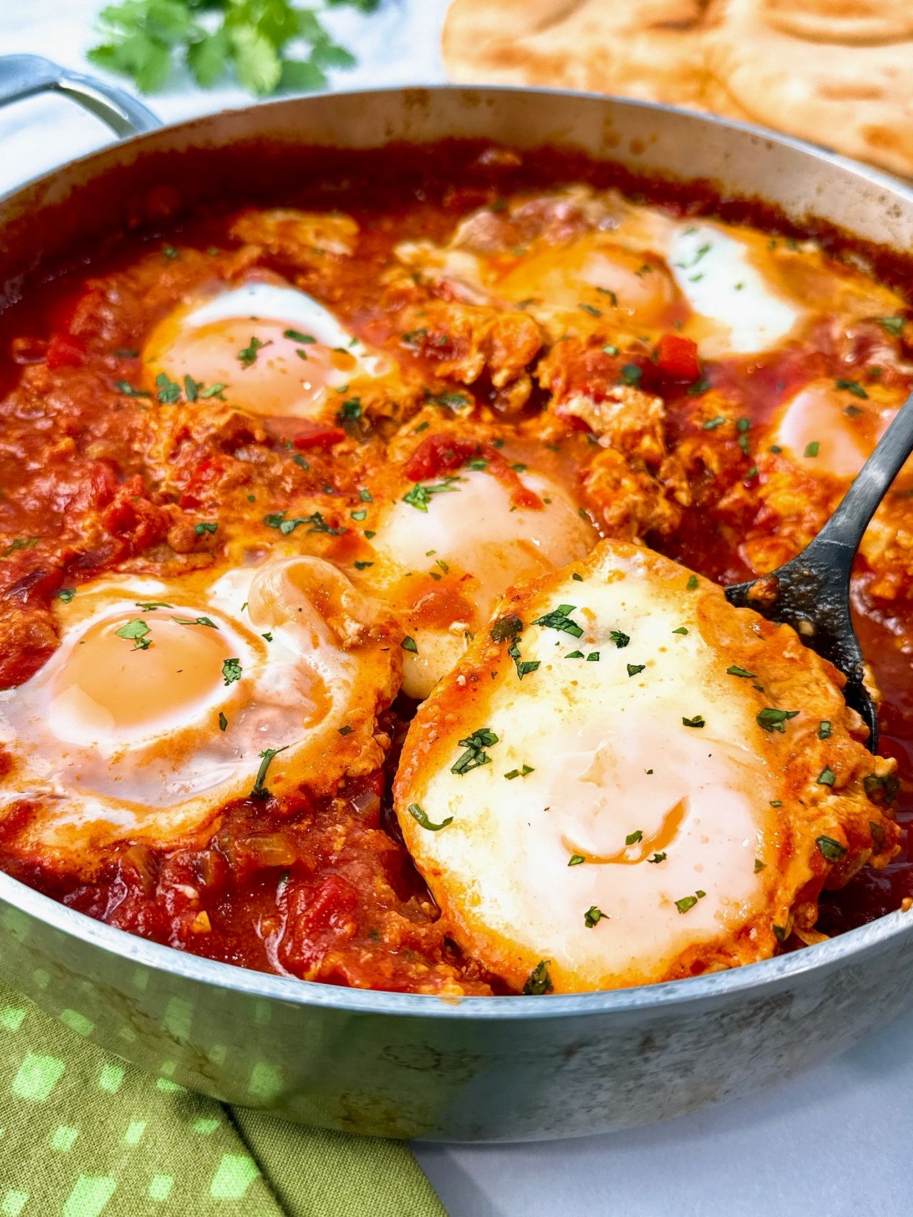 shakshuka in a silver pan with black spoon, green towels, and naan