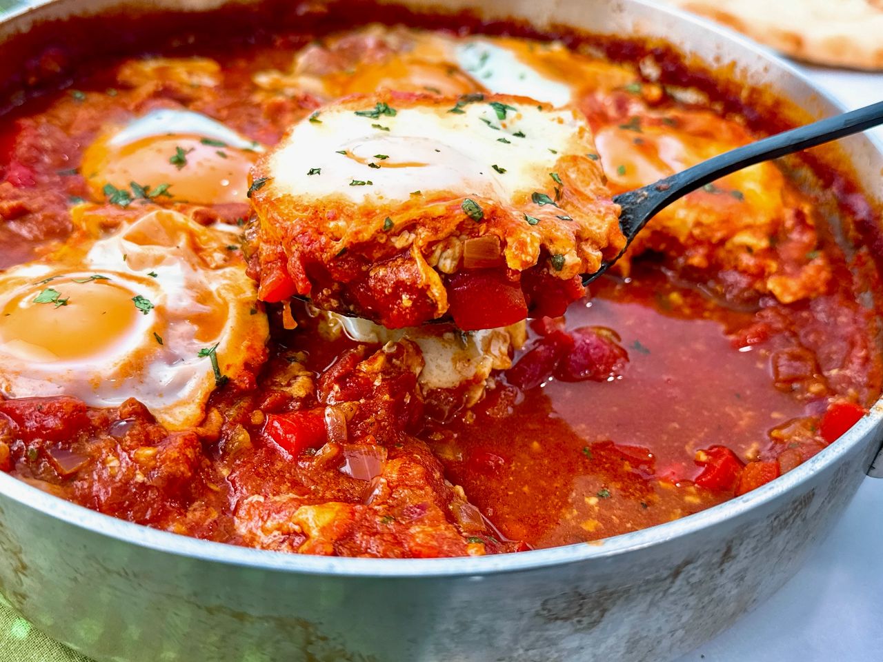 shakshuka in a silver pan with black spoon, green towels, and naan