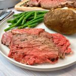 slices of prime rib on a white plate with a baked potato and green beans