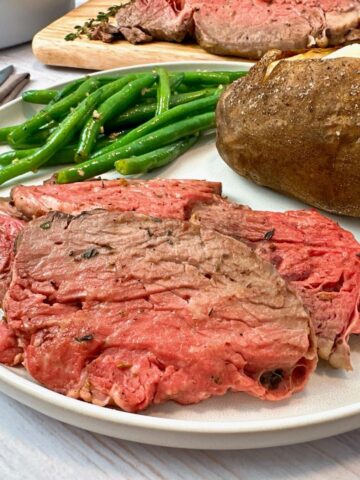 slices of prime rib on a white plate with a baked potato and green beans