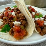 ground beef and bean soft tacos on a white plate with crumbled cheese garnish