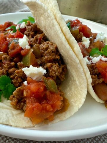 ground beef and bean soft tacos on a white plate with crumbled cheese garnish