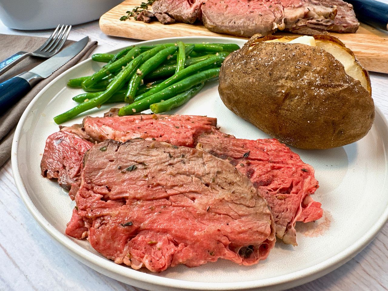 slices of prime rib on a white plate with a baked potato and green beans
