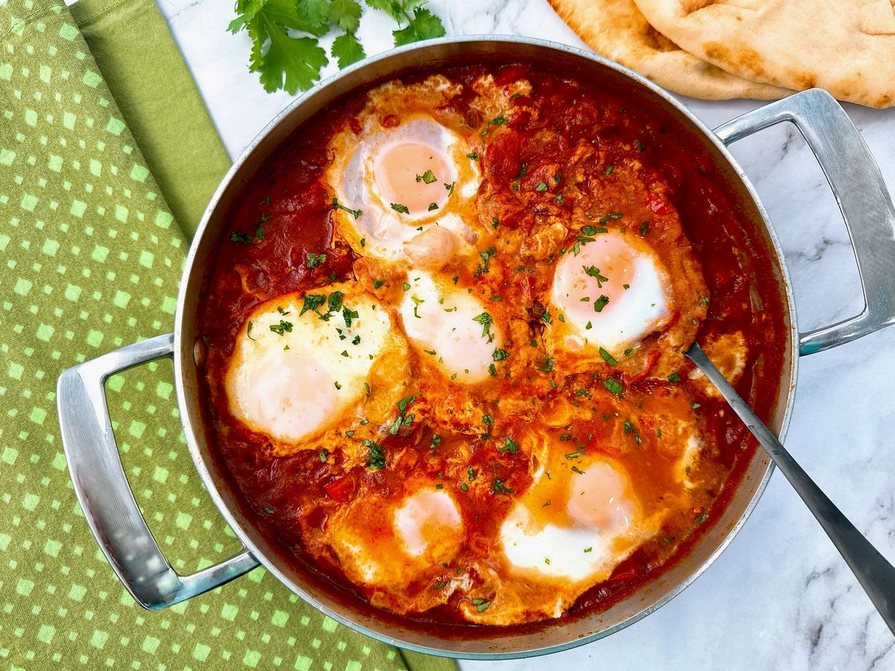 shakshuka in a silver pan with black spoon, green towels, and naan