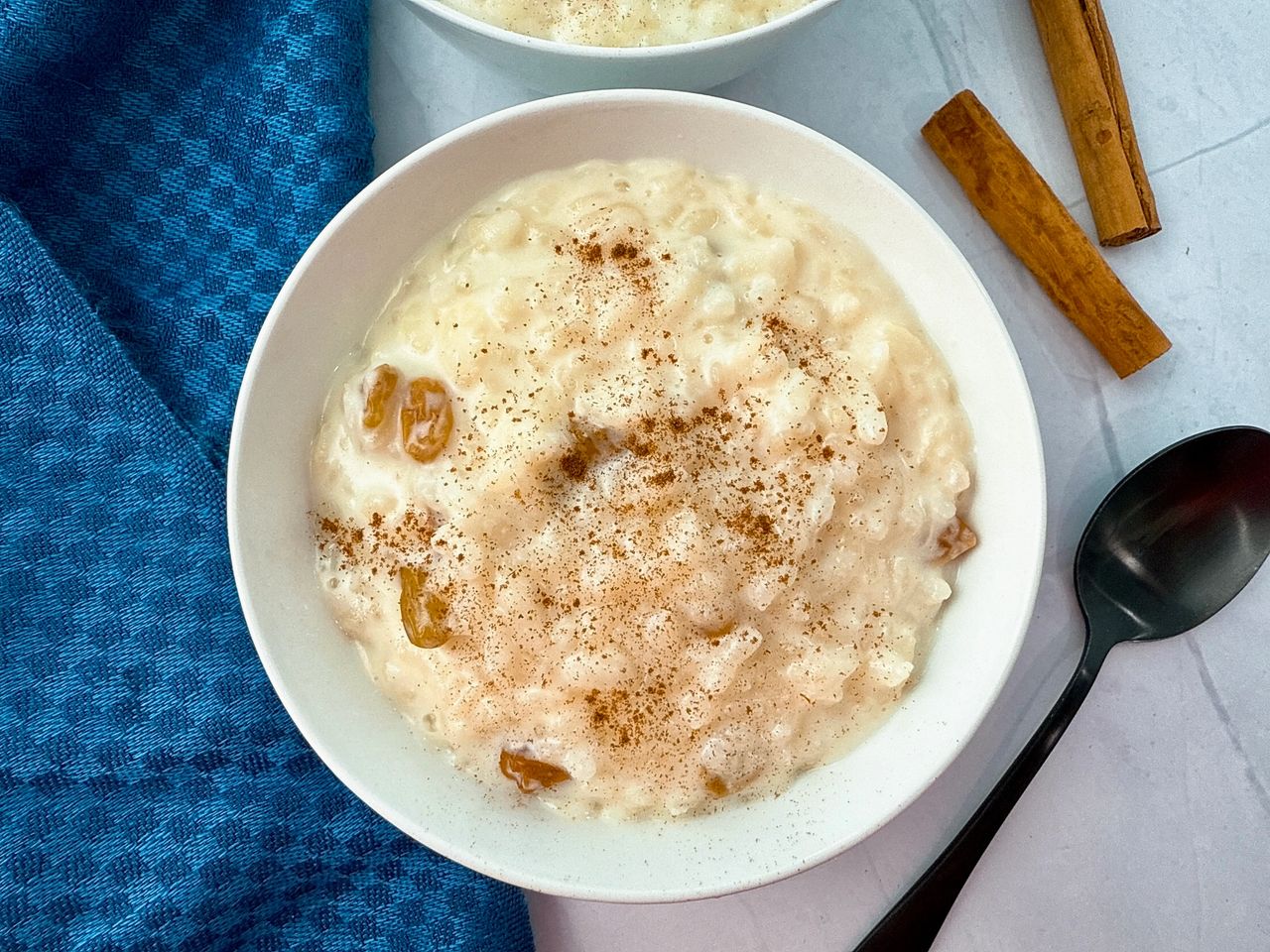 rice pudding in a white bowl with a black spoon and cinnamon sticks on the side