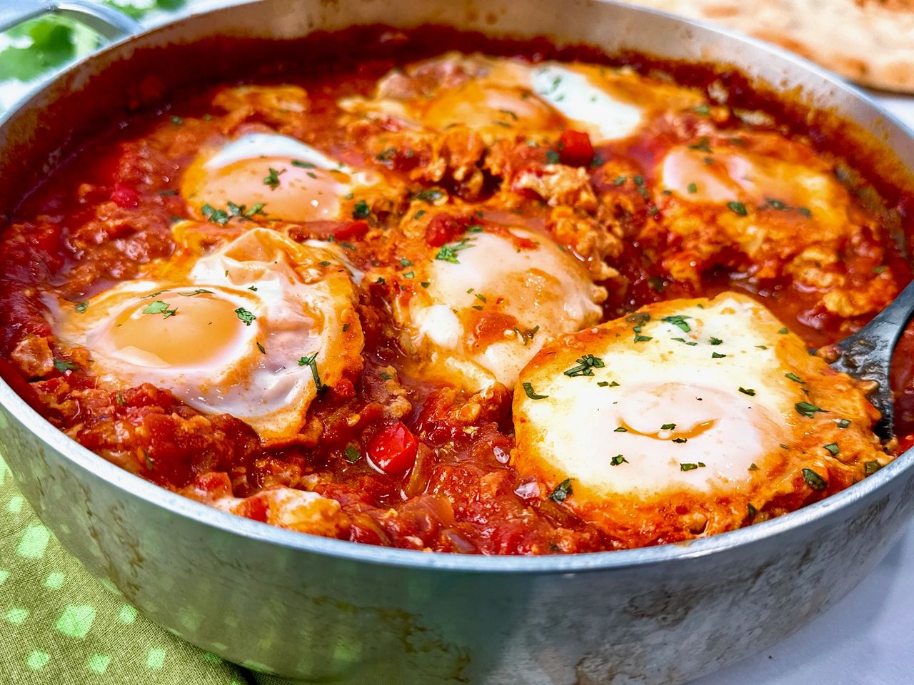 shakshuka in a silver pan with black spoon, green towels, and naan
