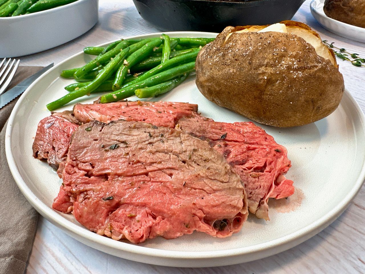 slices of prime rib on a white plate with a baked potato and green beans