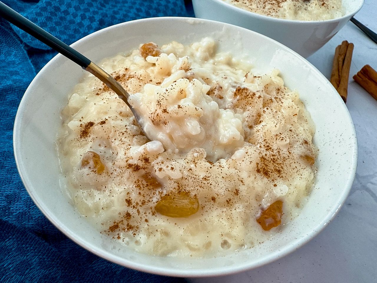 rice pudding in a white bowl with a black spoon and cinnamon sticks on the side