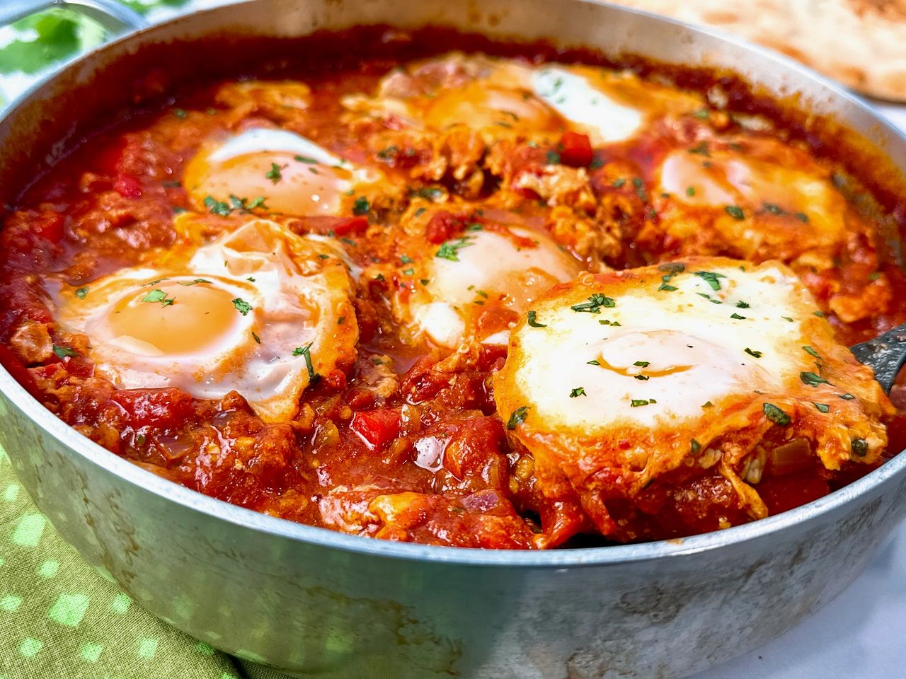 shakshuka in a silver pan with black spoon, green towels, and naan