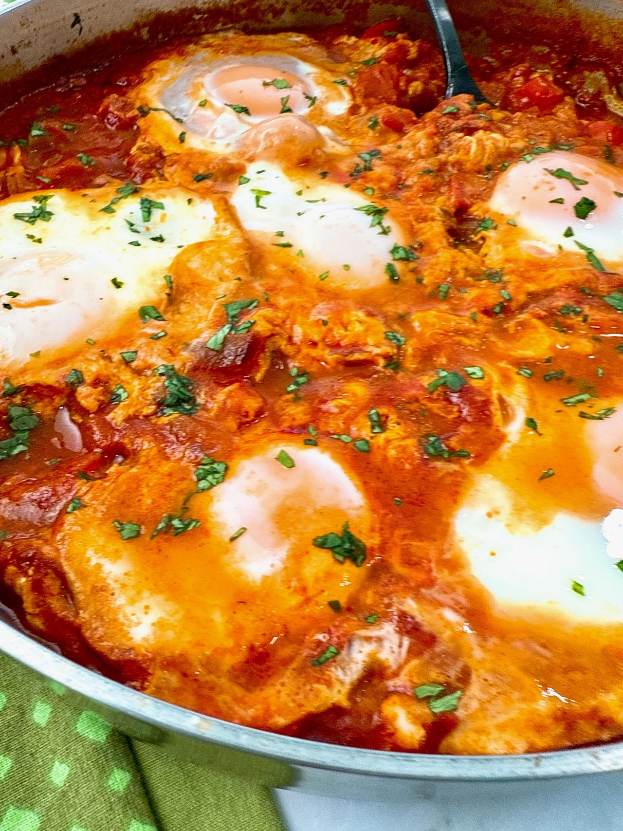 shakshuka in a silver pan with black spoon, green towels, and naan