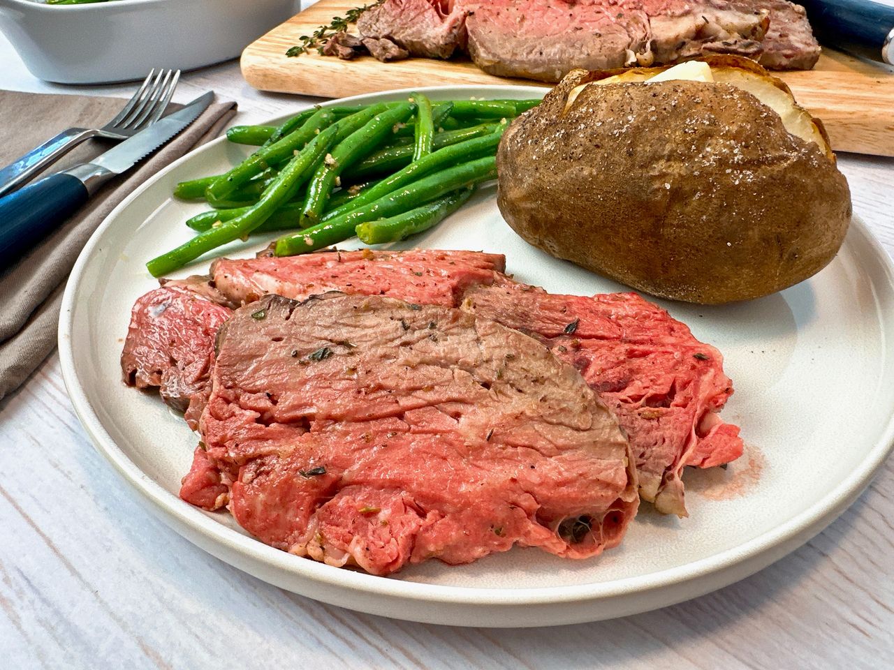 slices of prime rib on a white plate with a baked potato and green beans