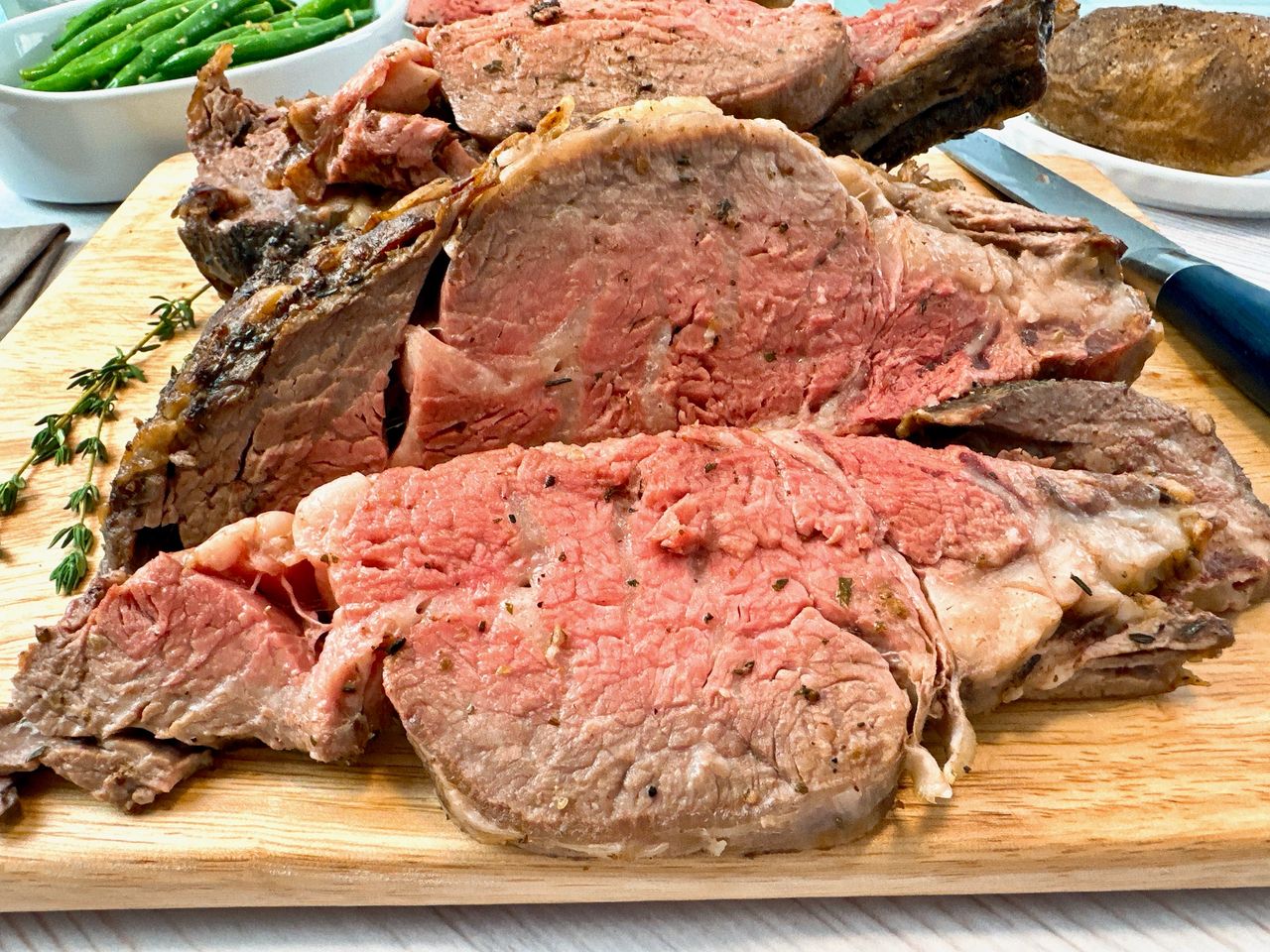 slices of prime rib on a cutting board with a baked potato and green beans