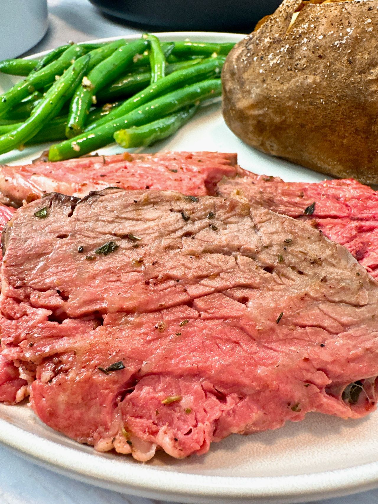 slices of prime rib on a white plate with a baked potato and green beans