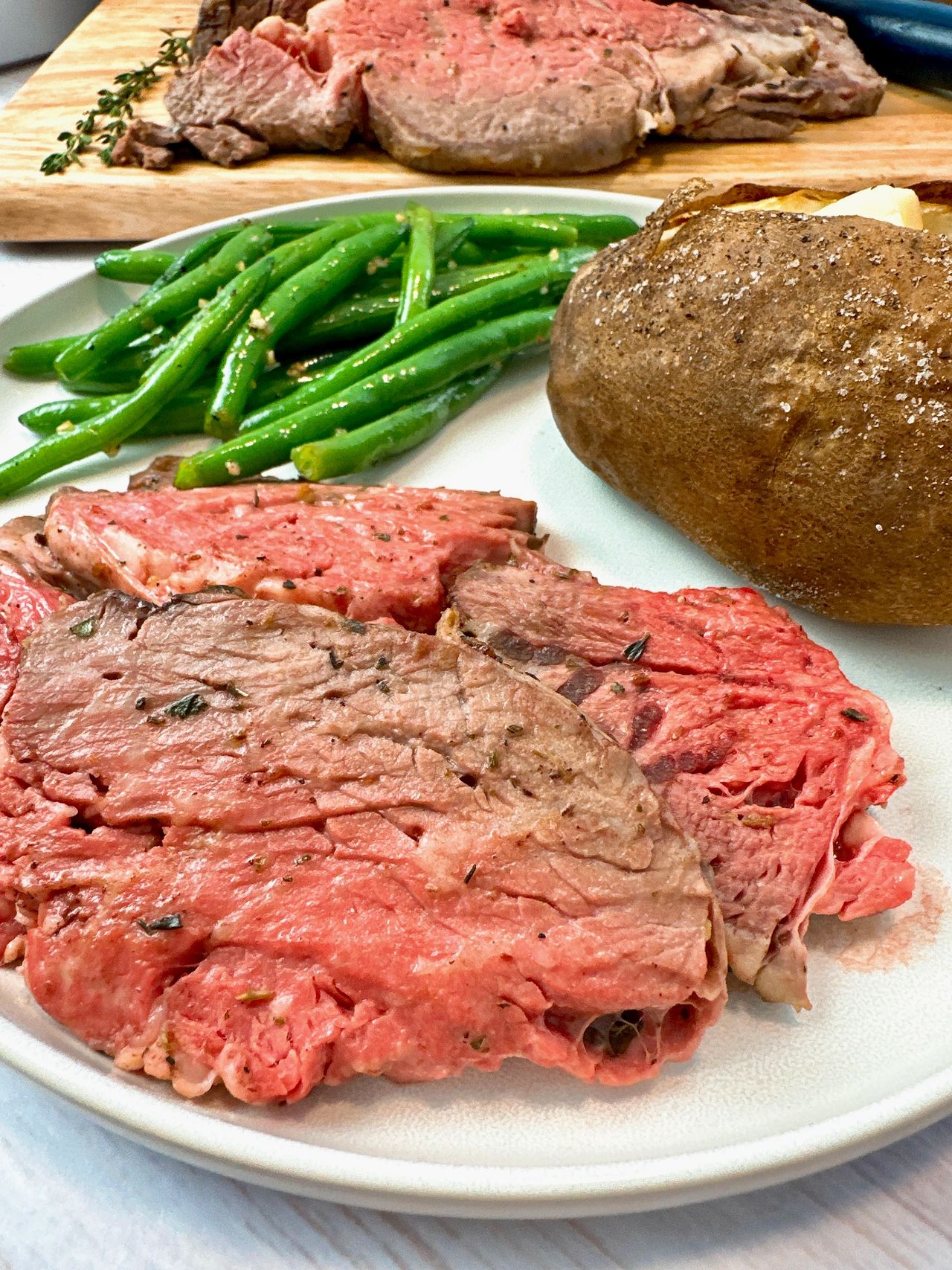 slices of prime rib on a white plate with a baked potato and green beans