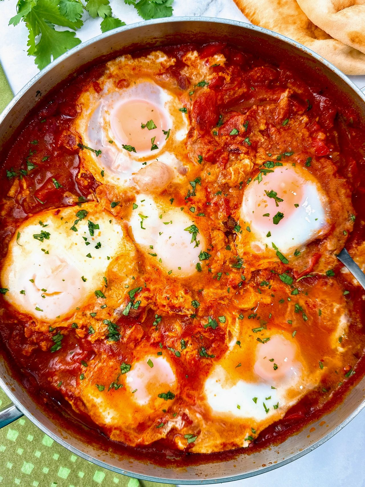 shakshuka in a silver pan with black spoon, green towels, and naan